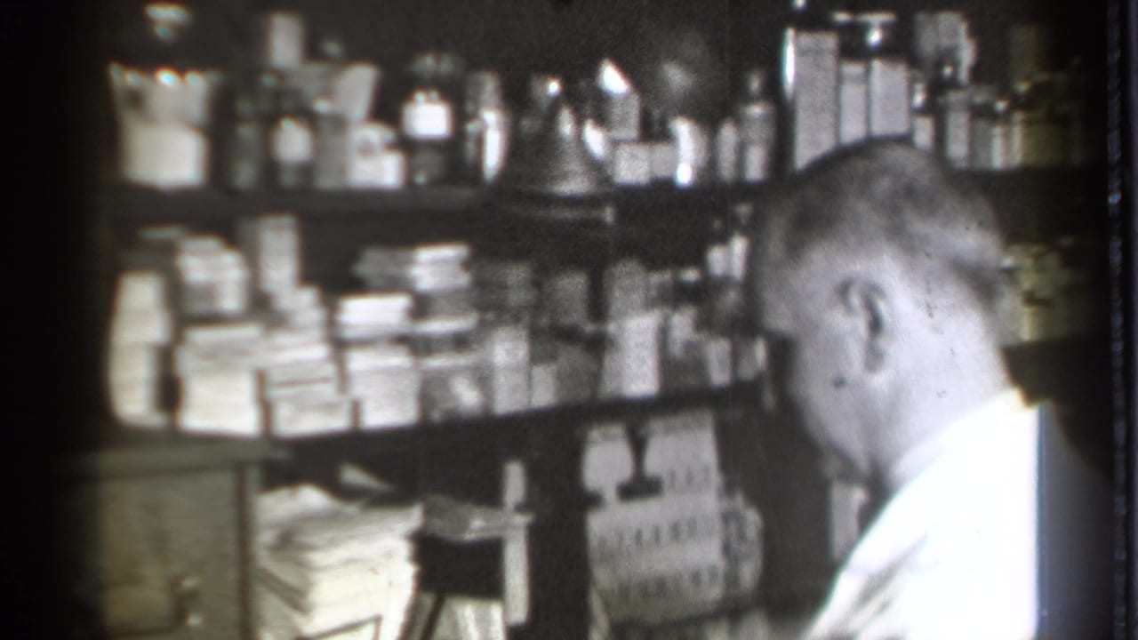 Pharmacist in a white coat seated beside shelves of bottles, boxes, and stacked papers.