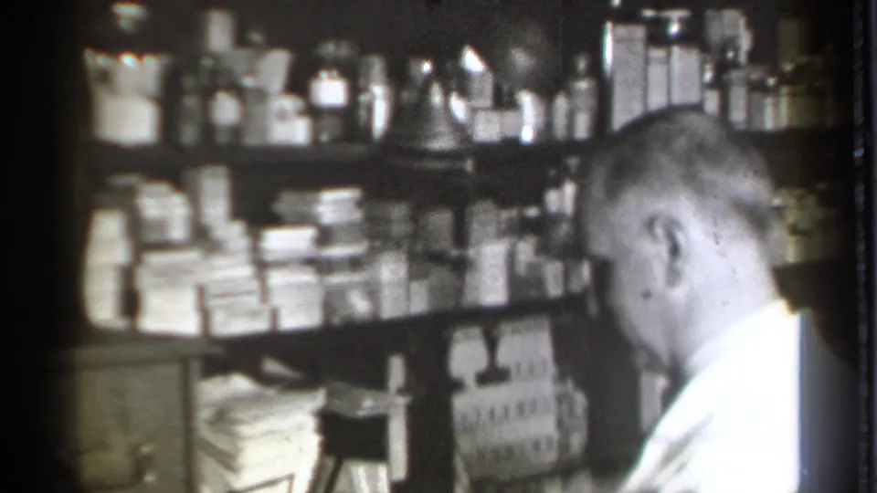 In a 1939 Danielson, Connecticut pharmacy interior, a white-coated pharmacist sits near shelves filled with bottles, boxes, and papers.