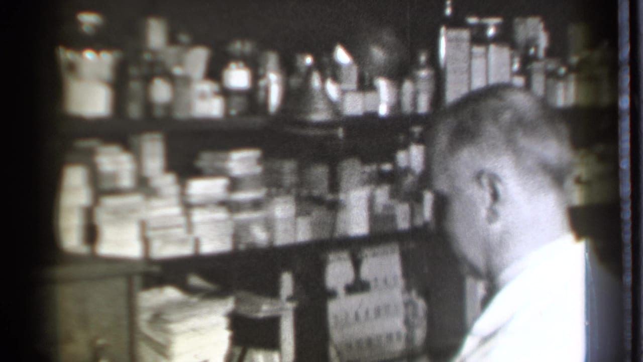 Side view of a pharmacist seated beside crowded shelves in a dim pharmacy interior.