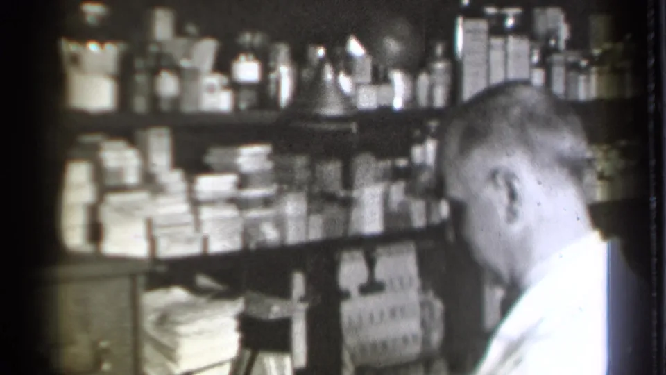 A closer 8mm view shows the pharmacist seated near densely packed shelves of pharmacy bottles, boxes, and paperwork.