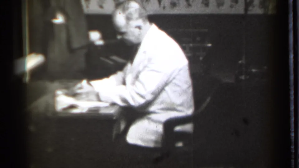 The pharmacist sits at a desk in the 1939 Danielson pharmacy, appearing to write or organize papers under dim interior light.