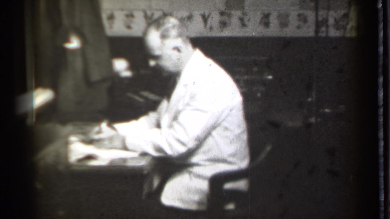 Man in a white coat sits at a desk writing or reviewing papers in a dim office.