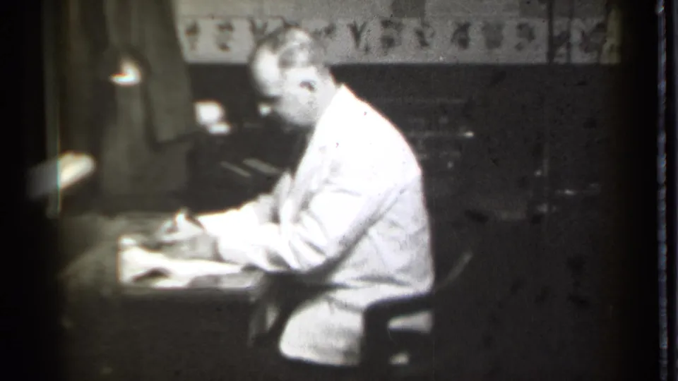 In 1939 Danielson, Connecticut footage, a man in a white coat leans over papers at a desk under a small lamp.