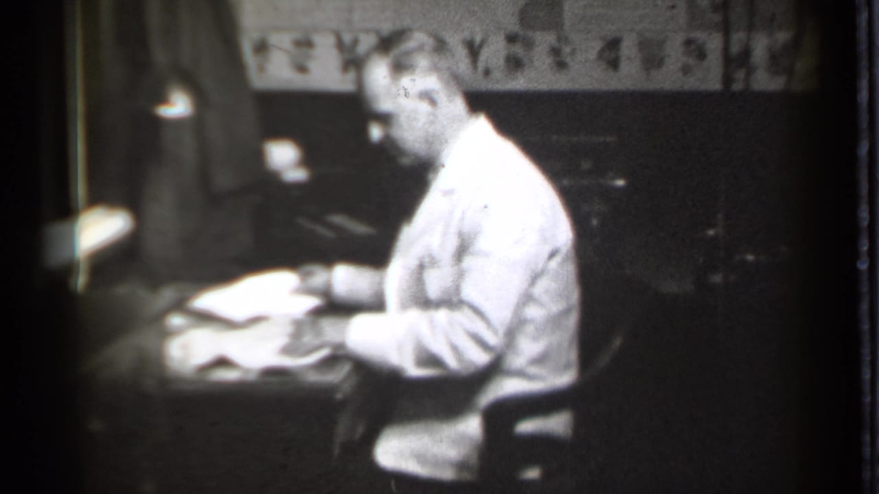 Seated man in white coat looks down at open papers on a desk.
