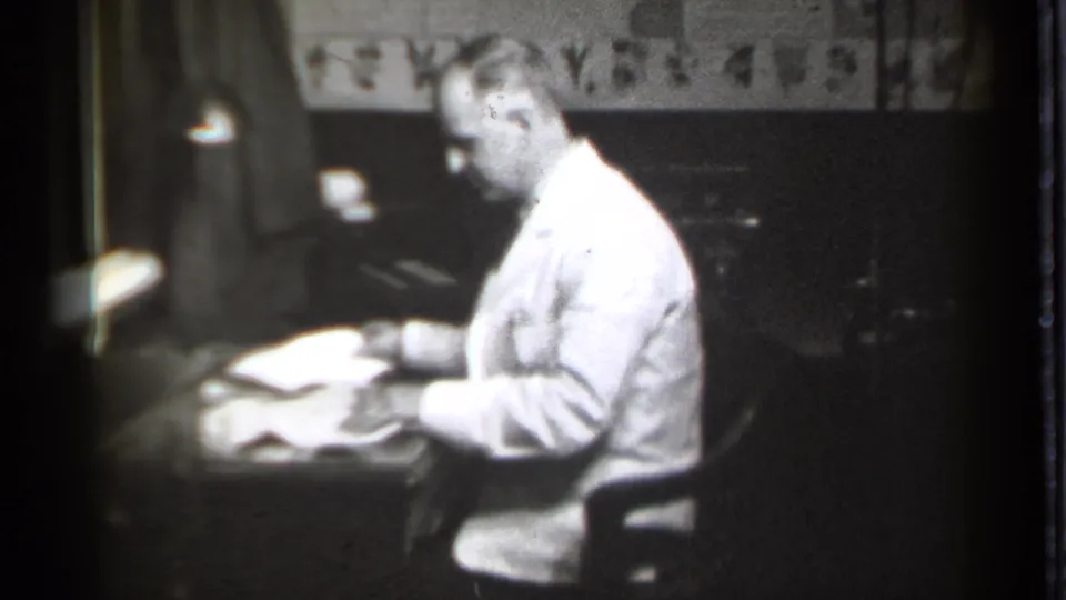 The seated man appears to review documents at a desk in Danielson, Connecticut, with a lamp and dark office furnishings nearby.