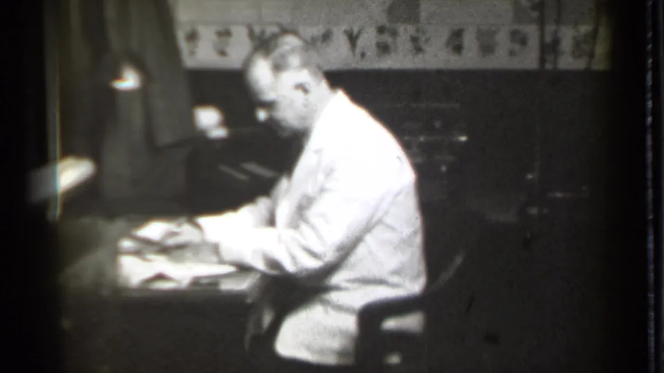 A man in a white coat bends toward documents on a desk, captured in grainy 8mm black-and-white footage from Danielson, Connecticut.