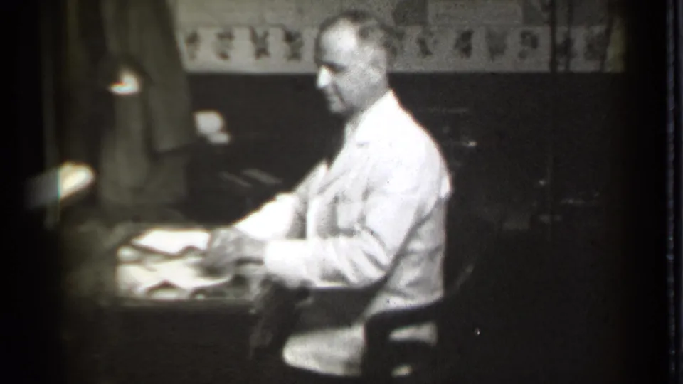 The man pauses or turns slightly toward the camera while seated at the desk, with papers, a lamp, and dark interior fixtures visible.