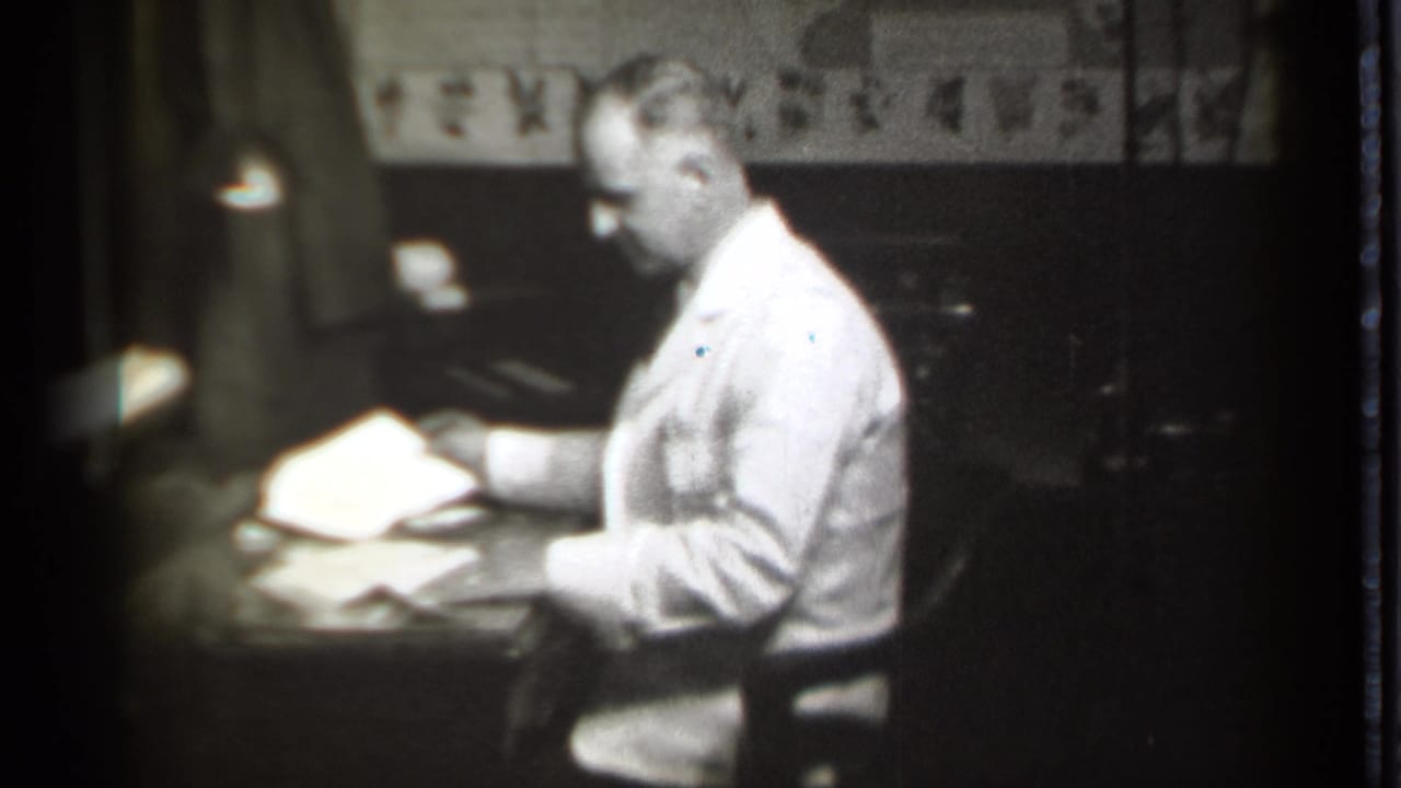 In Danielson, Connecticut, 1939 footage, a seated man in a light suit handles papers at a desk in a dark office.