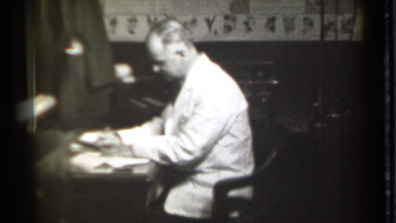 Seated man leans over papers and writes at a desk.