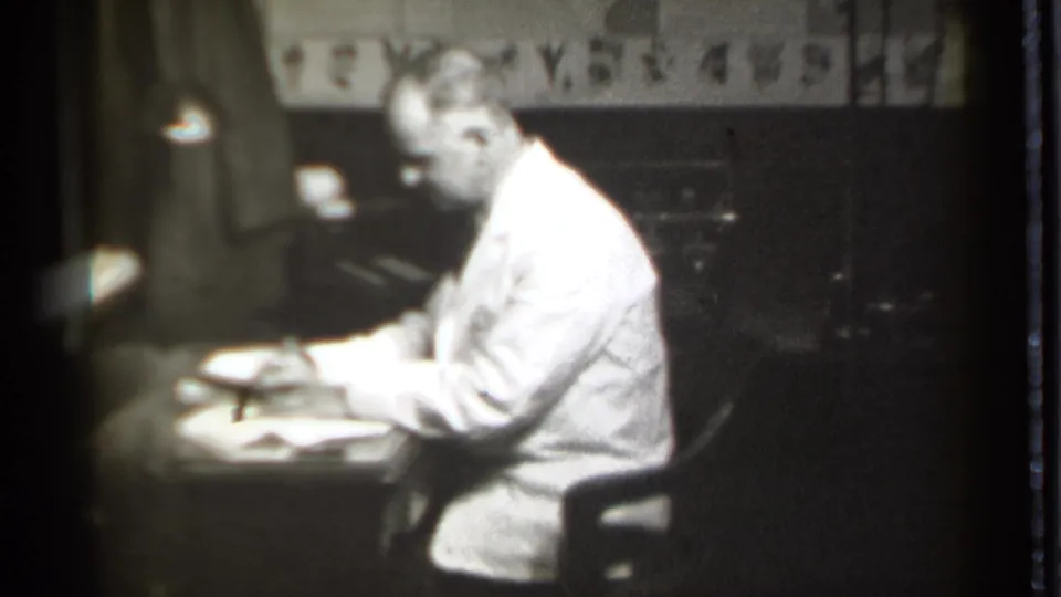 A man in a light suit continues writing at a desk, with loose documents and a lamp visible in the dim office setting.