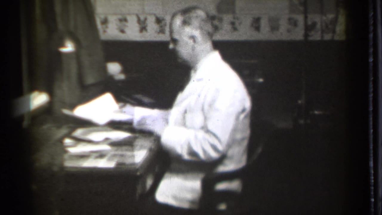 Man in a white coat sits at a desk and handles papers under a small lamp.