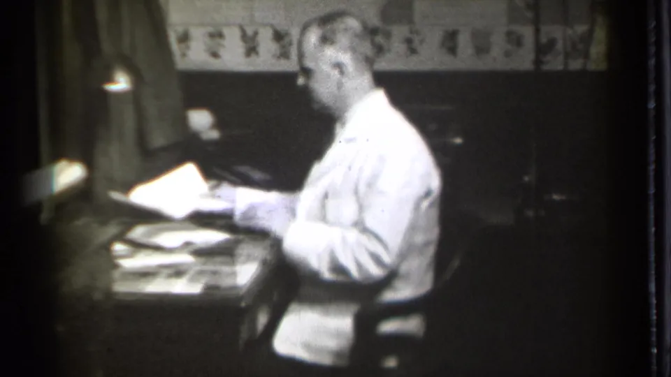 In Danielson, Connecticut in 1939, a man in a white coat sits at a desk with papers beneath a shaded lamp in a dim room.
