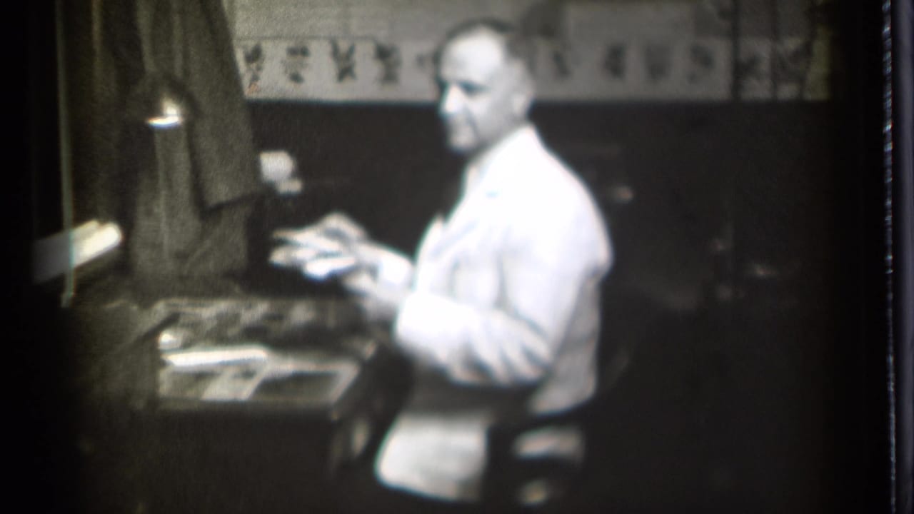 Man in a white coat faces partly toward the camera while seated at a desk.