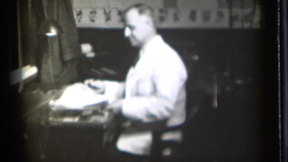 A man in a white coat works over papers at a desk in the Danielson, Connecticut interior, with a lamp and wall trim visible behind him.