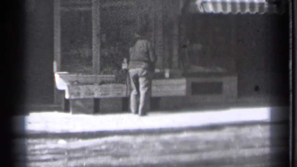 The man remains at the sidewalk counter in Danielson, Connecticut, with shop windows and display surfaces visible in soft 8mm focus.