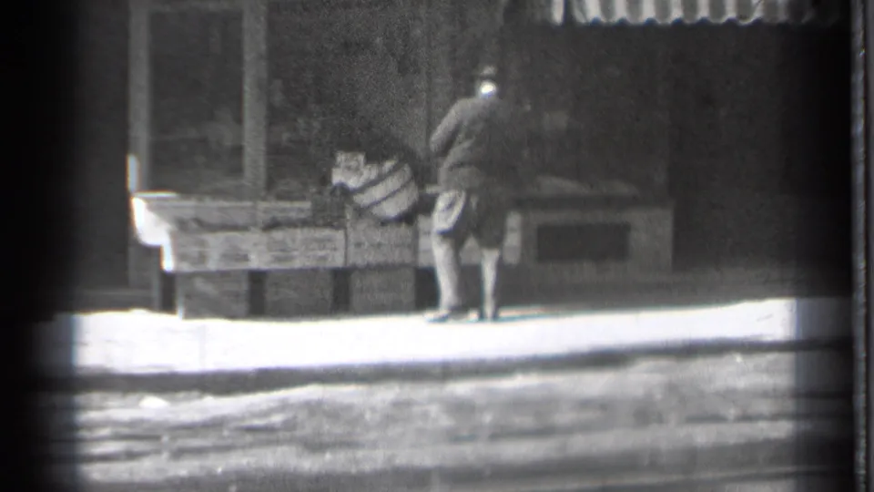 In Danielson, Connecticut, a man in a dark coat and hat bends beside wooden market crates on a sunny sidewalk in 1939 home-movie footage.