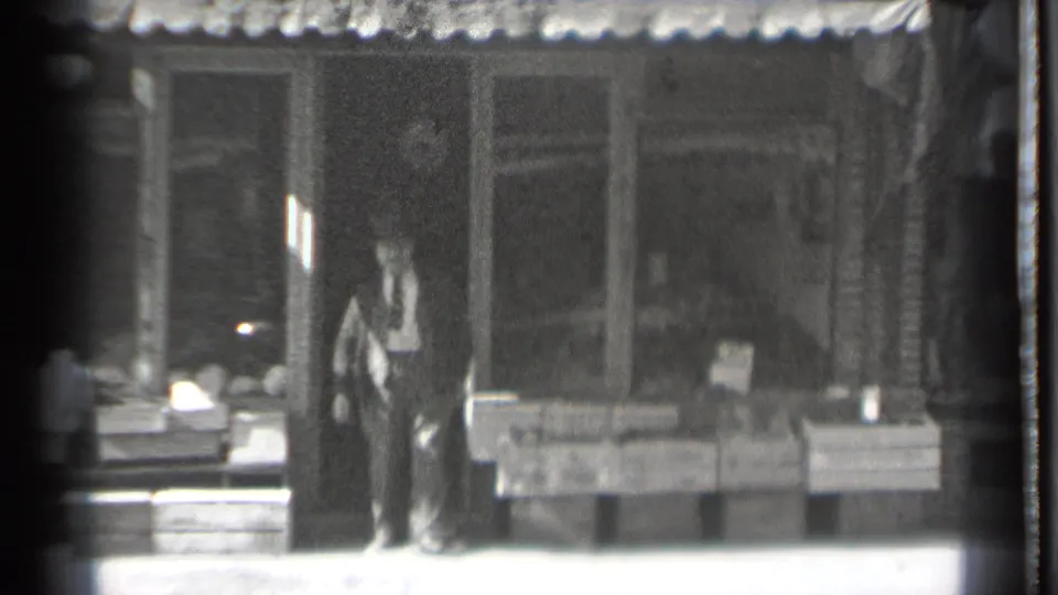 A suited man in a hat pauses near wooden crates and a storefront entrance in the Danielson, Connecticut market-front scene.