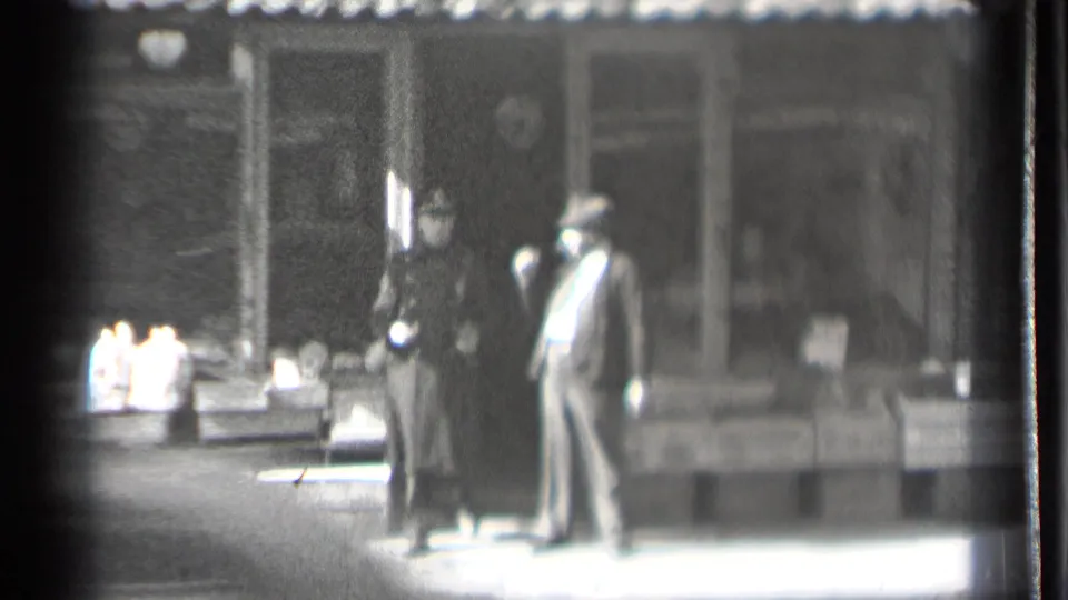 A man in a suit and hat appears to speak with a woman in a long coat near storefront display crates in Danielson, Connecticut.