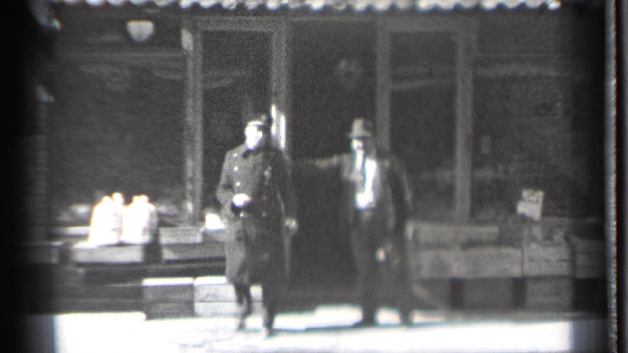 Two adult men stand outside a storefront, one in a dark uniform and one in a suit and hat.