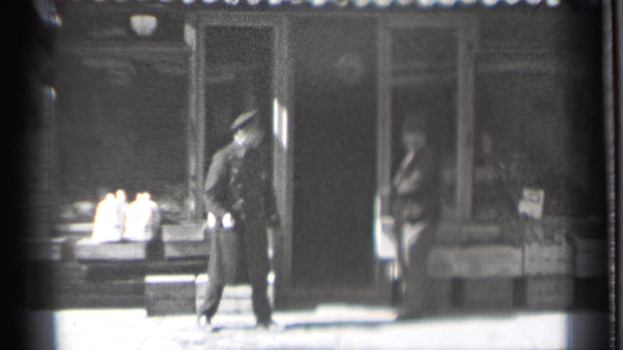 A uniformed man stands on the sidewalk while another man is partly visible in the storefront doorway.