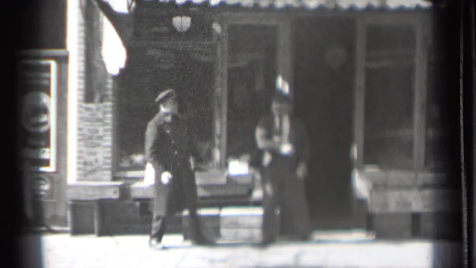 At the end of the Danielson sidewalk clip, the uniformed man remains by the storefront while a blurred adult figure crosses near the camera.