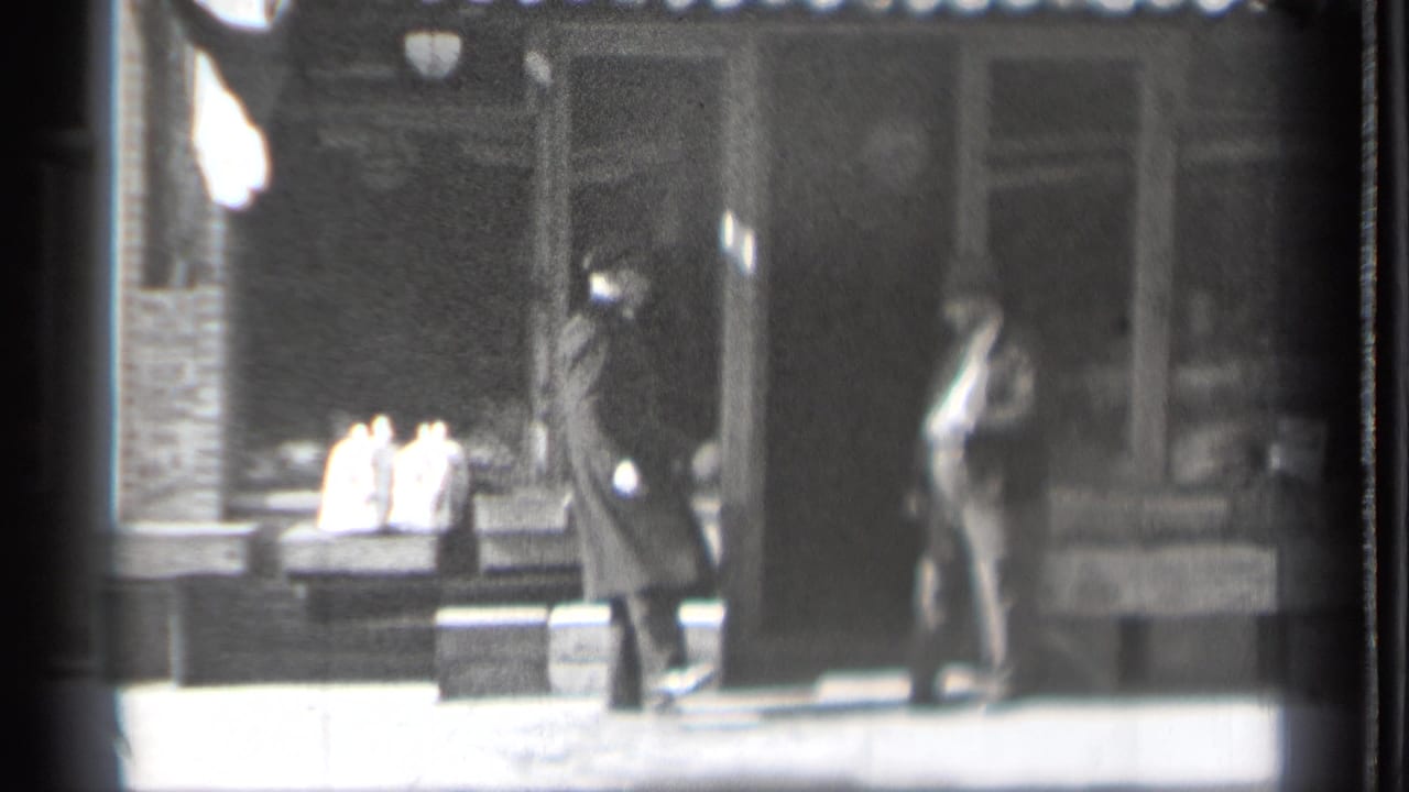 A dark-coated man and a lighter-suited man stand near a storefront entrance.