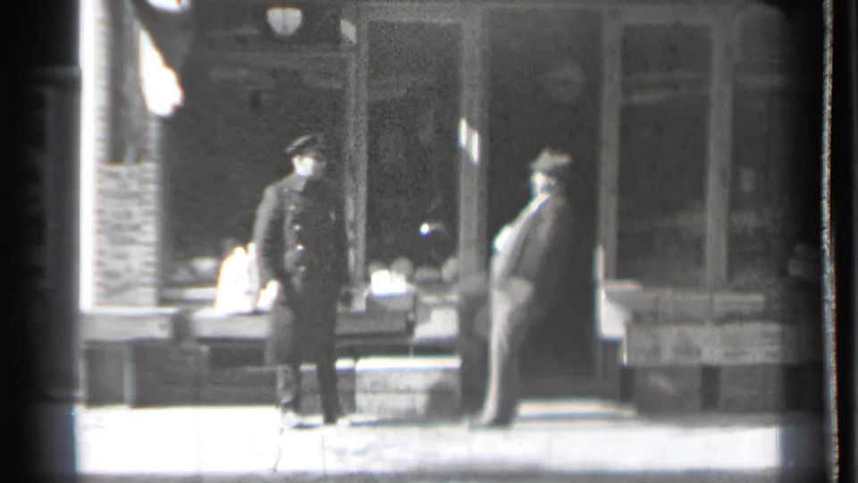 A dark-coated man and a heavier-set man in a lighter suit appear to pause in conversation outside a Danielson storefront in the supplied 1939 footage.