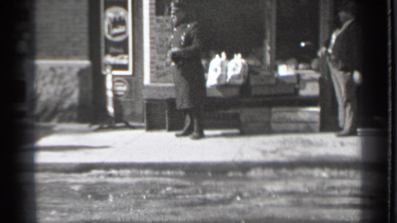 Pedestrians stand and walk near a storefront window on a small-town sidewalk.