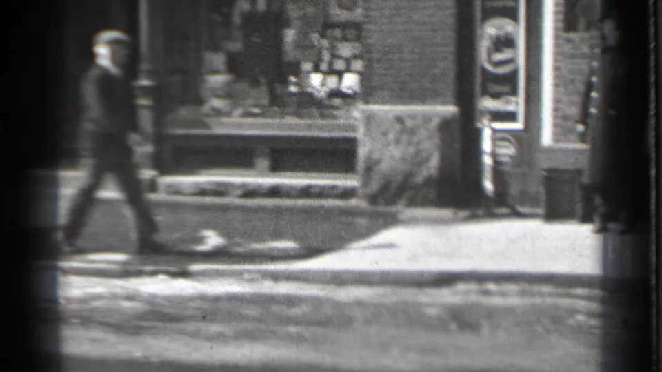 A man in period street clothing walks past a Danielson storefront window in the supplied 1939 home-movie footage.