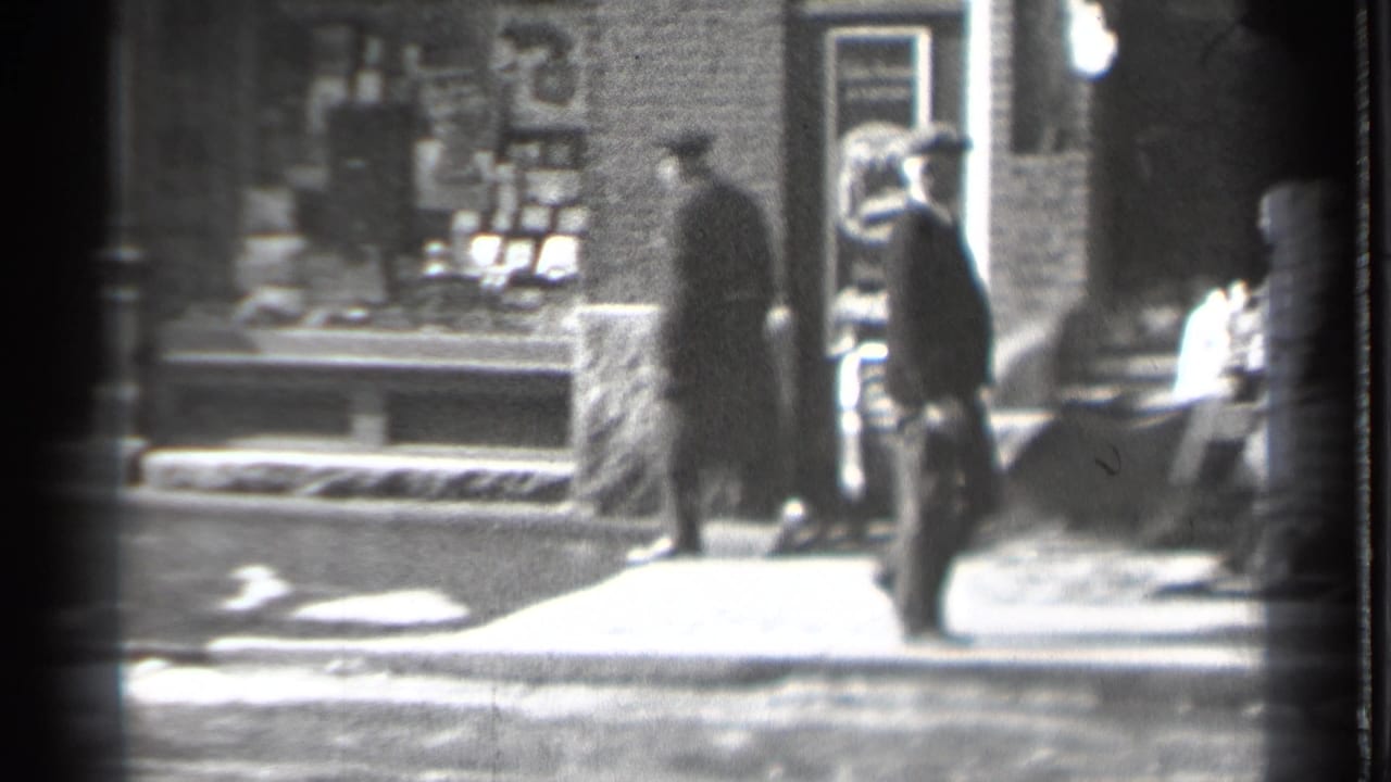 Pedestrians in coats pass brick storefronts and sidewalk displays on a winter street.