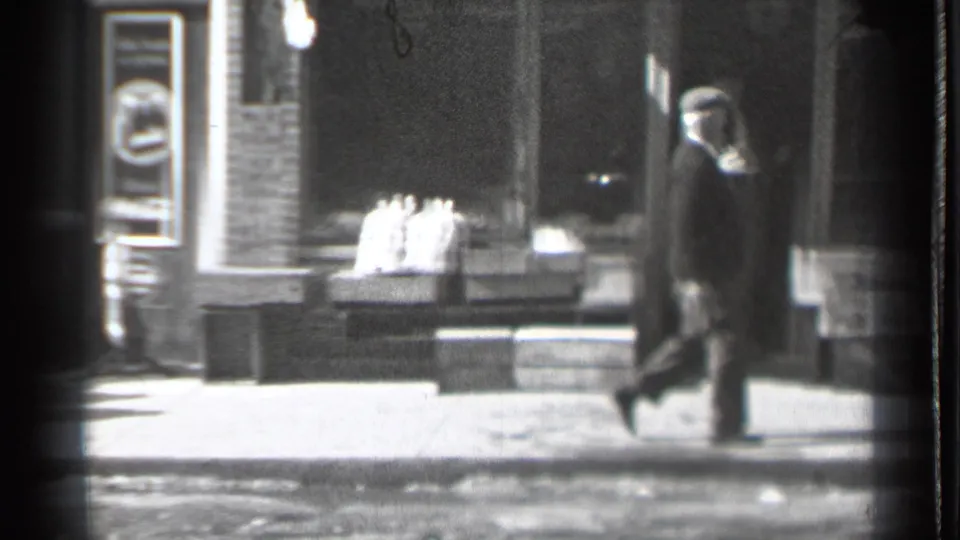 A 1939 Danielson sidewalk frame shows a coat-wearing pedestrian walking past stacked goods and brick shopfronts.