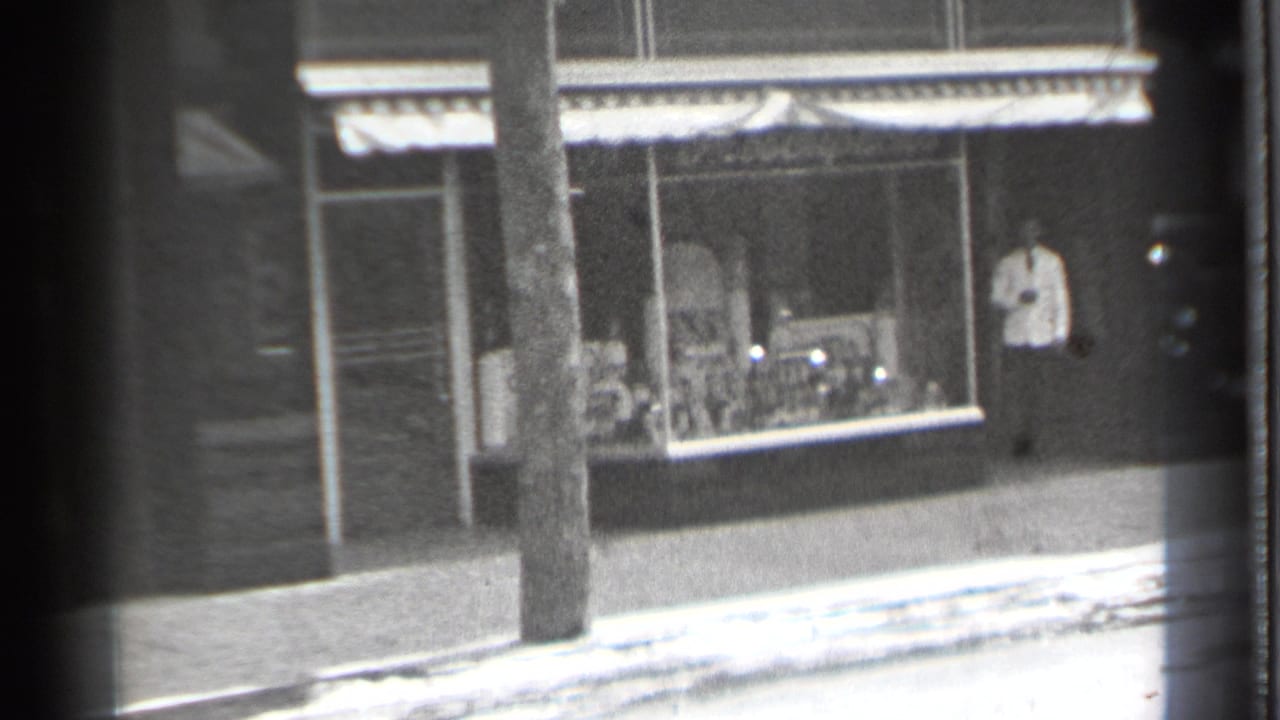 1939 Danielson, Connecticut storefront seen from a moving 8mm camera, with a man standing near the doorway and a utility pole in front.