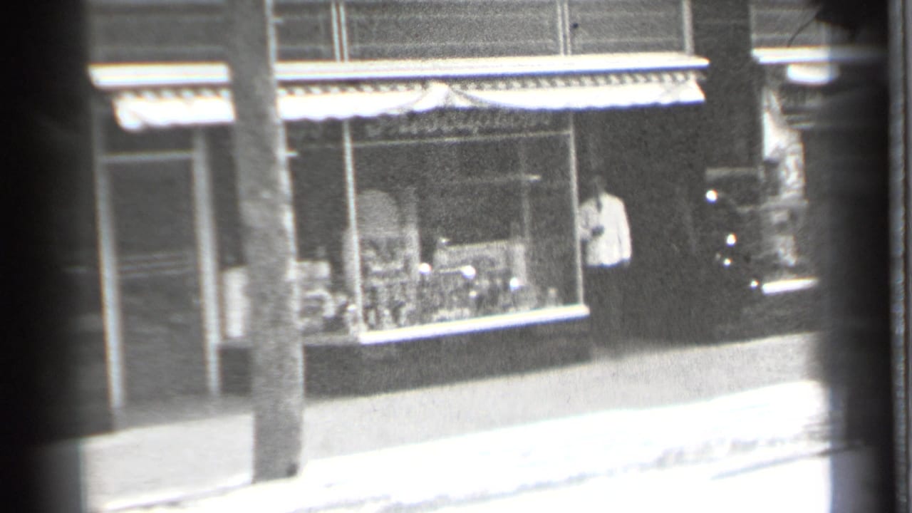 Street-facing storefront with awning, display windows, doorway figure, pole, and shaded building front.