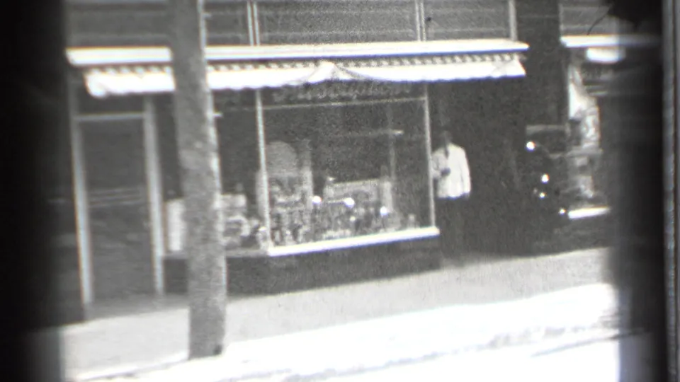 Danielson storefront with striped awning and a man near the entrance, filmed in soft black-and-white 8mm from across the street.