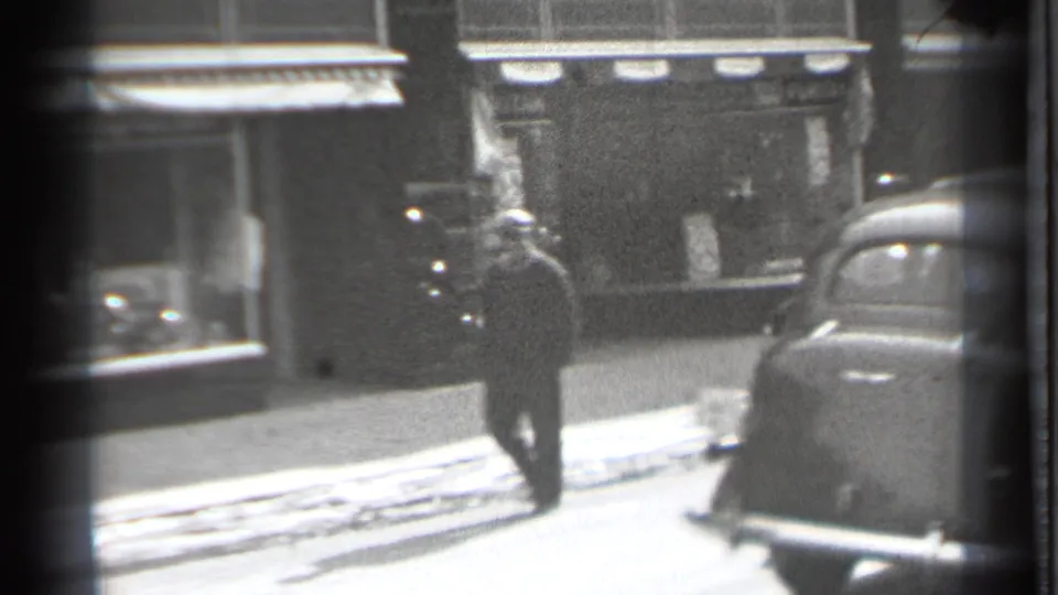 A pedestrian in dark clothing walks across the Danielson street scene, with storefront windows behind and a rounded automobile at frame right.