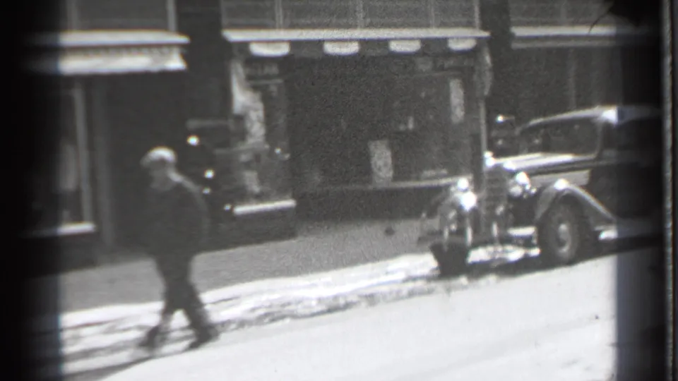 In Danielson, Connecticut, a pedestrian in dark clothing passes storefronts near a parked 1930s sedan on a snowy street.