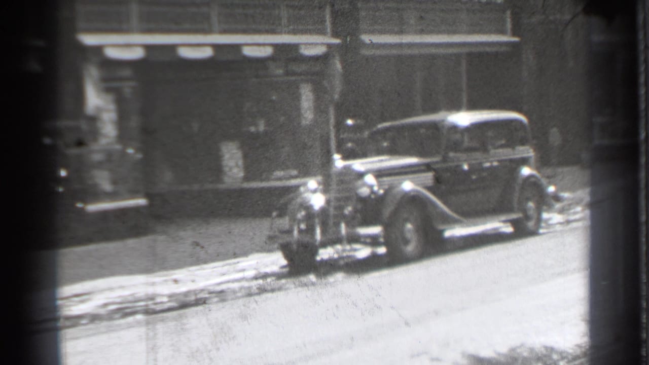 Parked old sedan beside storefronts with snow along the curb.