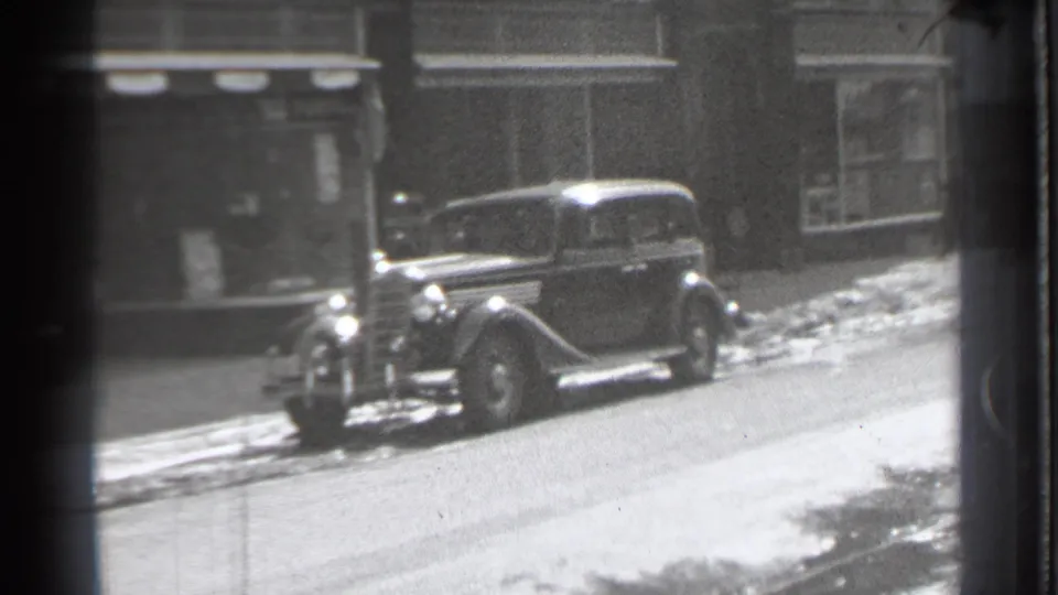The camera holds on a parked 1930s sedan along a snowy Danielson street, with storefront windows and awnings in the background.