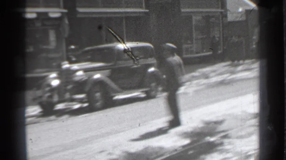 A pedestrian moves through the foreground as a parked 1930s sedan and snowy storefront street remain visible in Danielson, Connecticut.