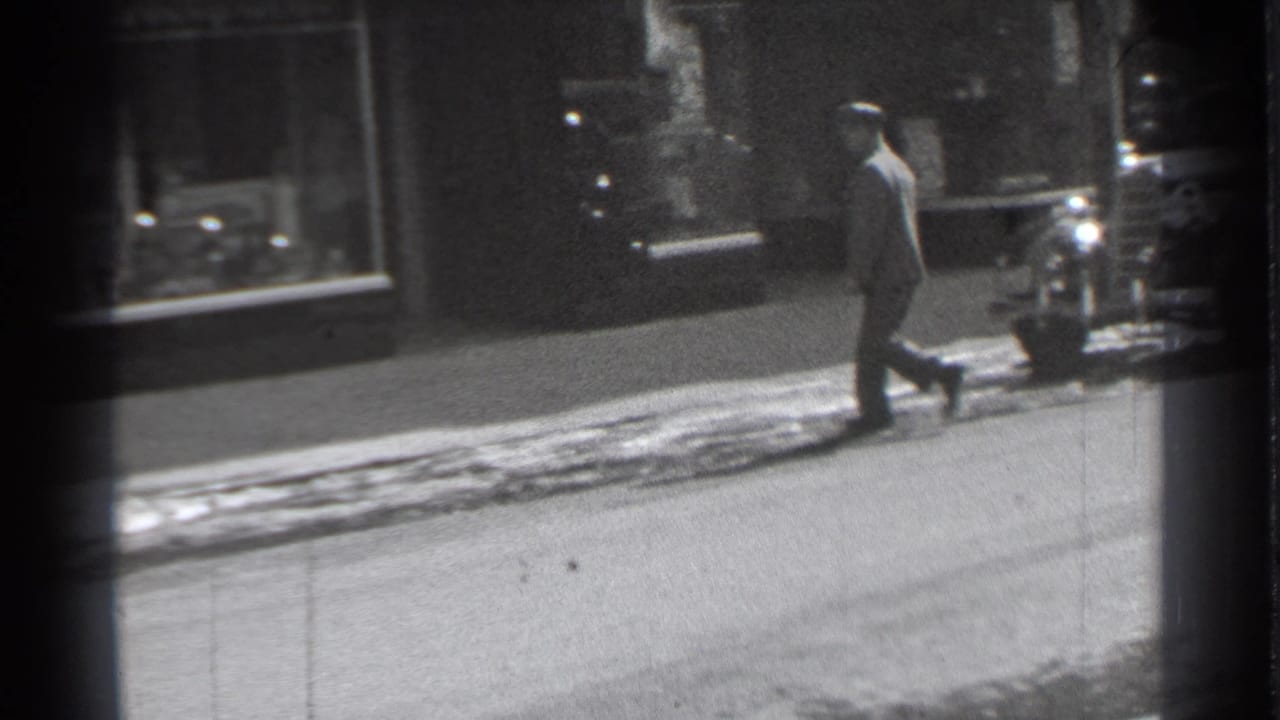 Man in a hat walks along a snowy sidewalk past storefront windows.