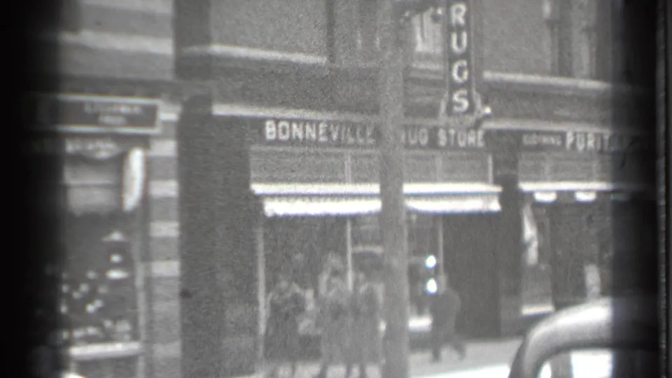 1939 Danielson commercial block with Bonneville Drug Store signage, a vertical RUGS sign, pedestrians, and a vintage car roof.