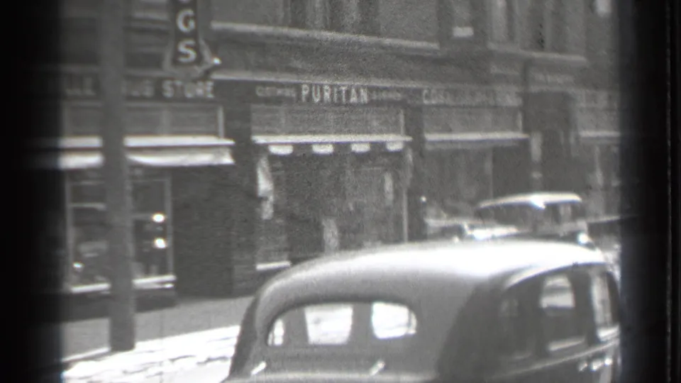 A 1939 Danielson downtown view centered near a Puritan storefront, with awnings, pedestrians, and cars visible through soft 8mm focus.