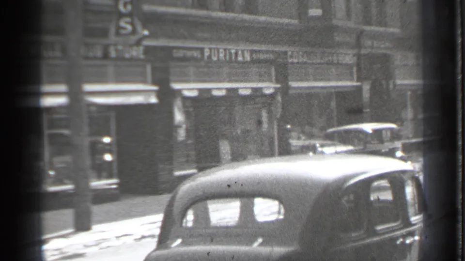1939 Danielson street view with parked rounded cars, storefronts, and a visible Puritan sign in soft black-and-white 8mm footage.
