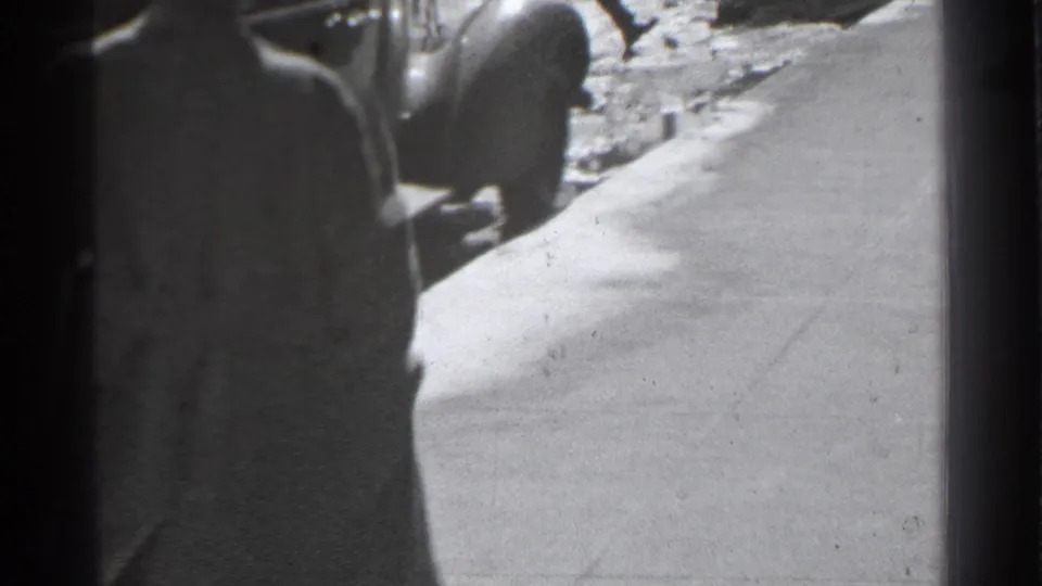 A man in a long coat walks along a Danielson sidewalk past a rounded parked car in 1939 black-and-white home-movie footage.