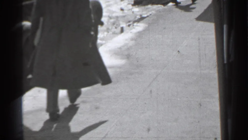 A long-coated pedestrian walks away along a bright Danielson sidewalk, with curbside snow or light debris visible ahead.