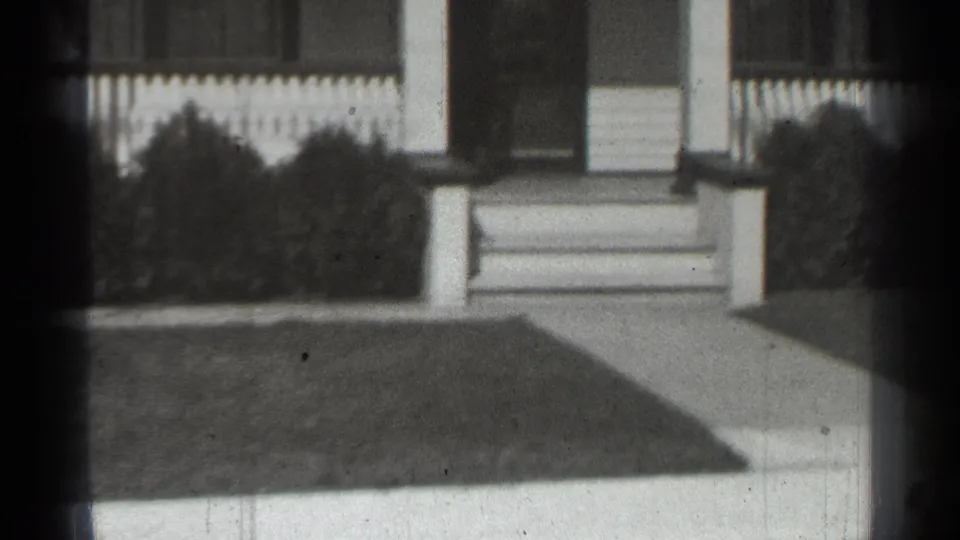 1939 New York 8mm view of a suburban house front with porch columns, steps, picket fence, shrubs, walkway, and lawn.