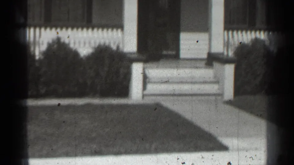 1939 New York home-movie frame showing a house entrance, porch supports, steps, picket fence, shrubs, walkway, and lawn.