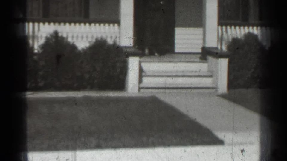 Soft 8mm frame of a 1939 New York suburban porch area with steps, column, picket fence, shrubs, walkway, and lawn.
