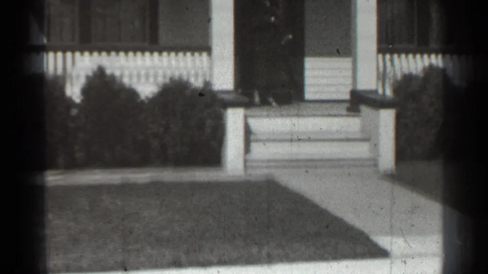 1939 New York frame of a suburban house front showing porch steps, fence, shrubs, walkway, lawn, and a dark doorway.