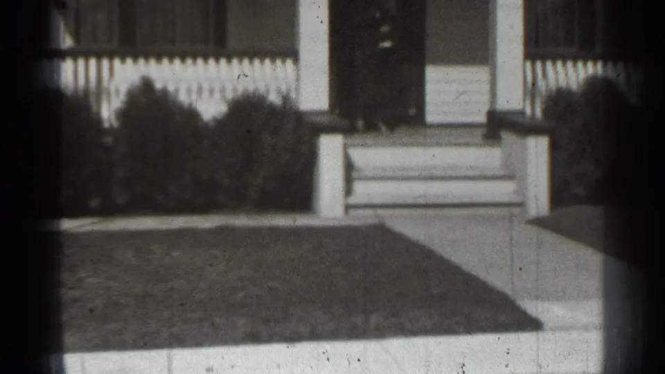 1939 New York home-movie frame showing a house porch, front steps, railings, shrubs, and lawn from a fixed outdoor viewpoint.